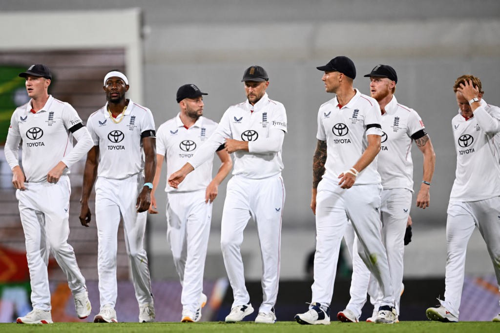 England cricket players walking together on the field during a Test match in white uniforms under stadium lights.
